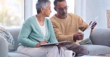 A senior couple sits together on a couch and looks at various papers. The woman has a clipboard on her lap.