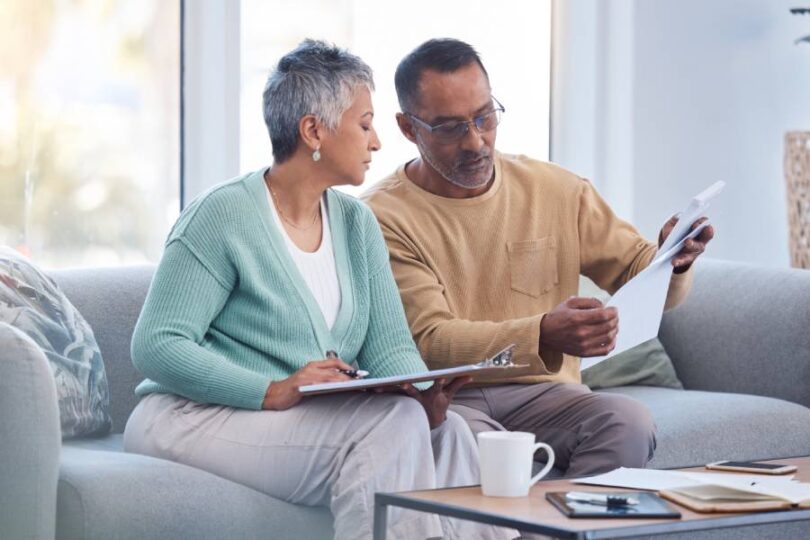 A senior couple sits together on a couch and looks at various papers. The woman has a clipboard on her lap.