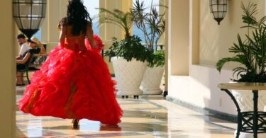 The back side of a girl in a poofy, red ball gown walking down a hall as two people sitting on a nearby bench look at her.