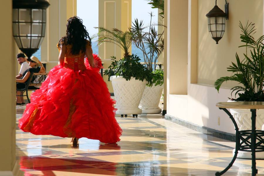 The back side of a girl in a poofy, red ball gown walking down a hall as two people sitting on a nearby bench look at her.