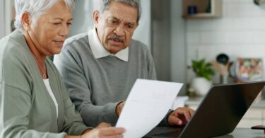 An older adult couple sits in their kitchen with a laptop in front of them. They look at a sheet of paper the woman is holding.