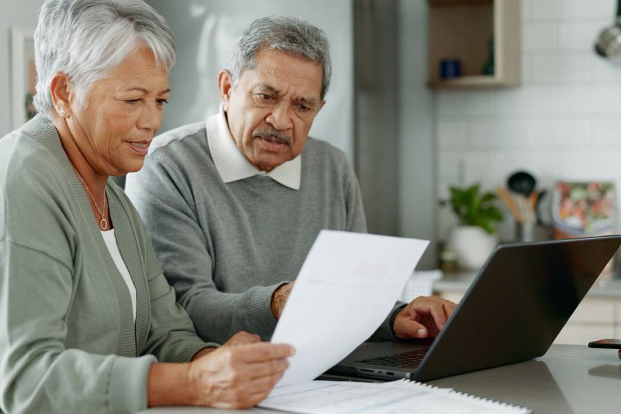 An older adult couple sits in their kitchen with a laptop in front of them. They look at a sheet of paper the woman is holding.