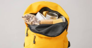 A yellow, unzipped backpack in front of a gray background. The backpack has concealed food items like cereal and crackers.