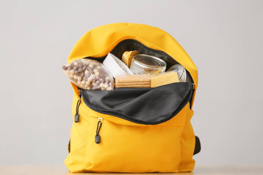 A yellow, unzipped backpack in front of a gray background. The backpack has concealed food items like cereal and crackers.