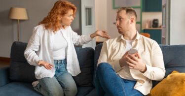 A couple on a couch. The woman is on her knees, gesturing angrily at the man, who is holding his phone and staring blankly at her.