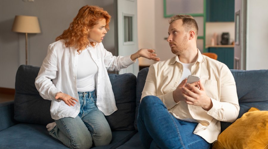 A couple on a couch. The woman is on her knees, gesturing angrily at the man, who is holding his phone and staring blankly at her.