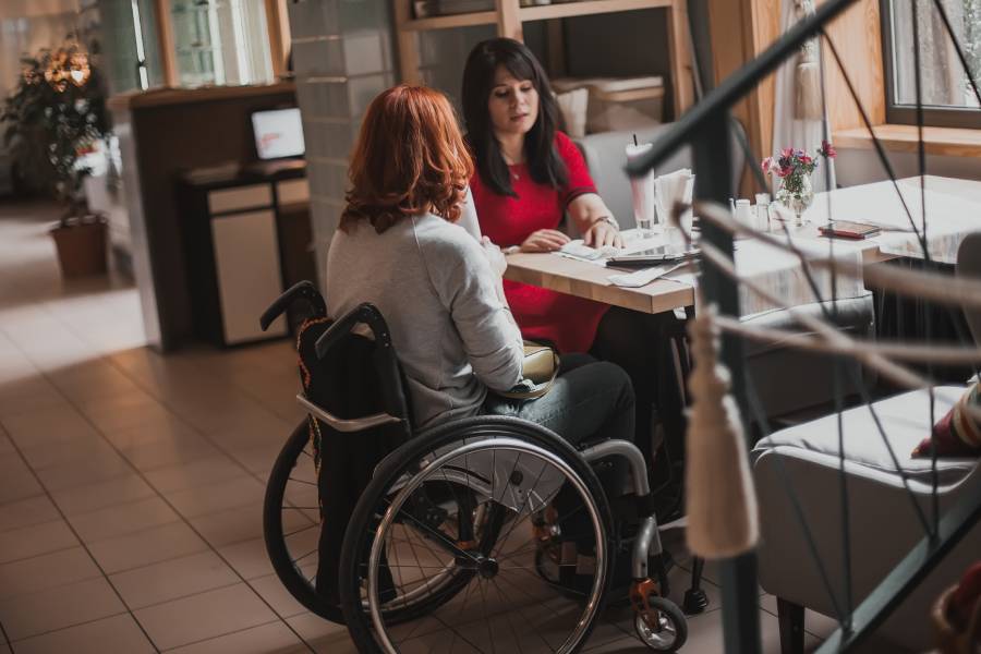 Two women sit at a restaurant table. One woman is a wheelchair user and sits at the end of the table.