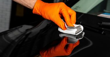 An individual wearing orange gloves is using a sponge and a microfiber cloth to polish a black vehicle.