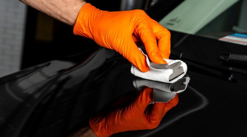 An individual wearing orange gloves is using a sponge and a microfiber cloth to polish a black vehicle.