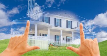 A close-up of female hands framing a half blueprint, half rendered image of a house set atop small, rolling green hills.
