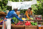 A woman grocer at a farmer's market prepares bins of fresh vegetables and fruits while talking to another grocer.