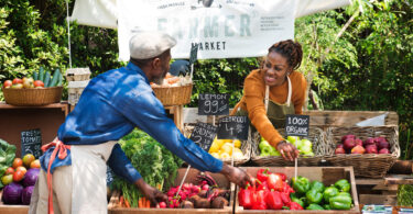 A woman grocer at a farmer's market prepares bins of fresh vegetables and fruits while talking to another grocer.
