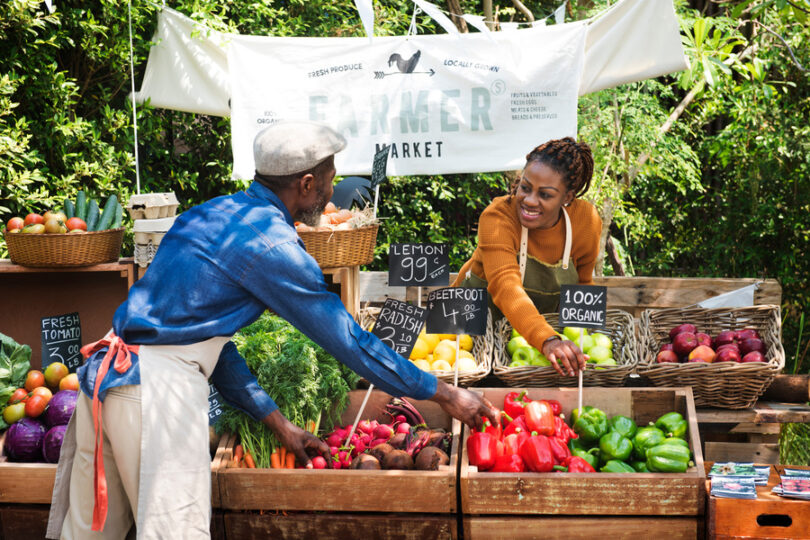 A woman grocer at a farmer's market prepares bins of fresh vegetables and fruits while talking to another grocer.