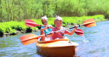 Two blonde boys paddle an orange canoe on a river on a sunny day. One is wearing sunglasses.