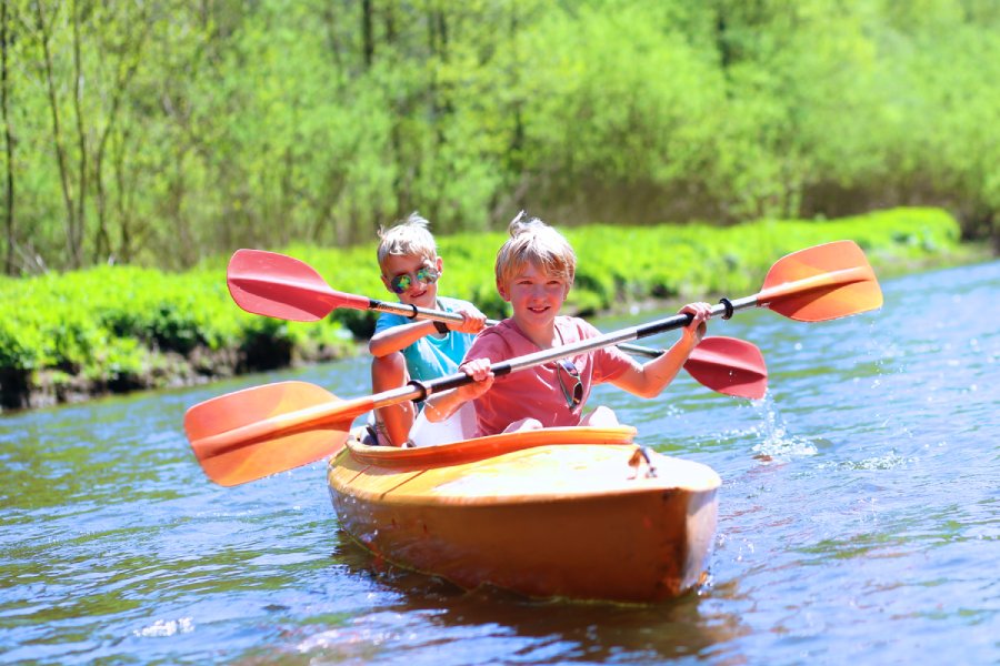 Two blonde boys paddle an orange canoe on a river on a sunny day. One is wearing sunglasses.