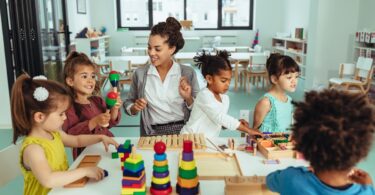 A group of children and a women gathered around a small table in a classroom setting for children as they play with colorful toys.