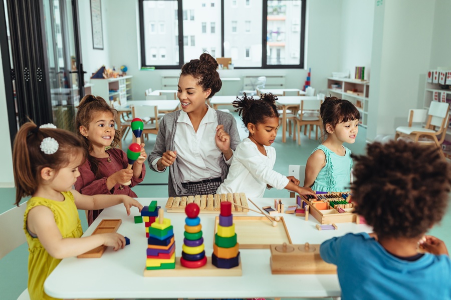 A group of children and a women gathered around a small table in a classroom setting for children as they play with colorful toys.