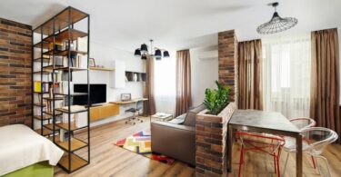 A fully furnished studio apartment with a bed, room dividers, a couch, and a dining table. There's brick and brown accents.