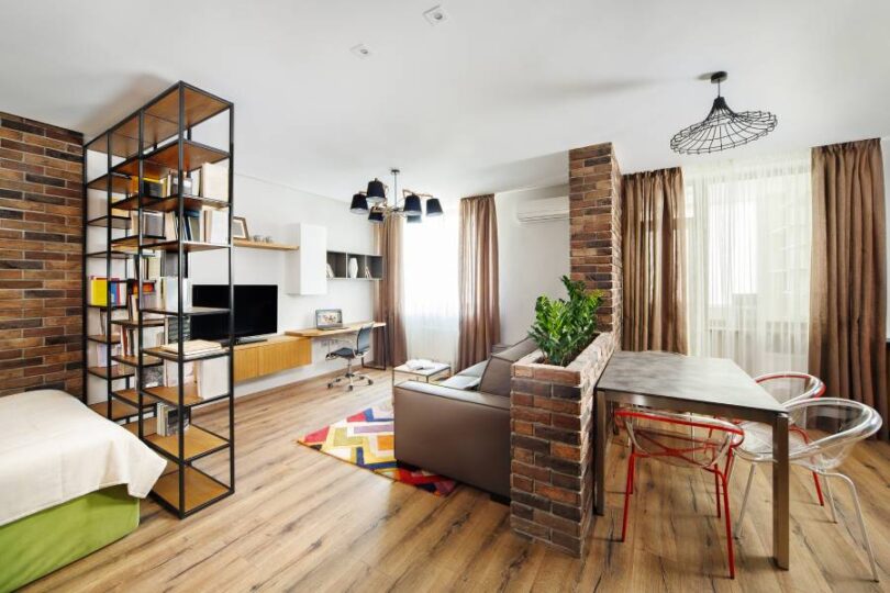 A fully furnished studio apartment with a bed, room dividers, a couch, and a dining table. There's brick and brown accents.