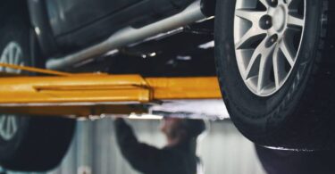 A closeup view of a yellow auto lift holding up a black car. In the background is a man in a jumpsuit working on the car.