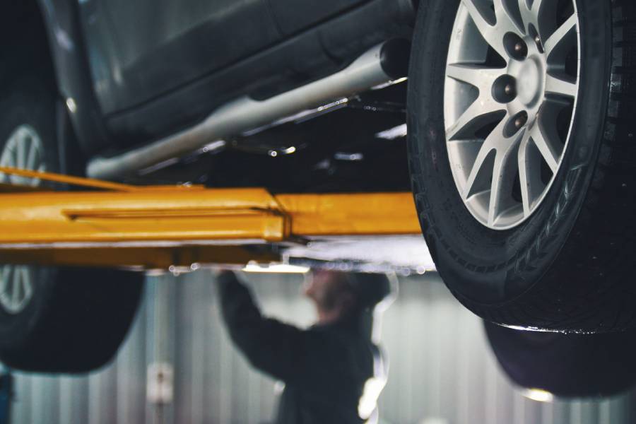 A closeup view of a yellow auto lift holding up a black car. In the background is a man in a jumpsuit working on the car.