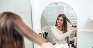 A woman with brown and blonde highlighted hair is smiling is straightening her hair in a salon chair.