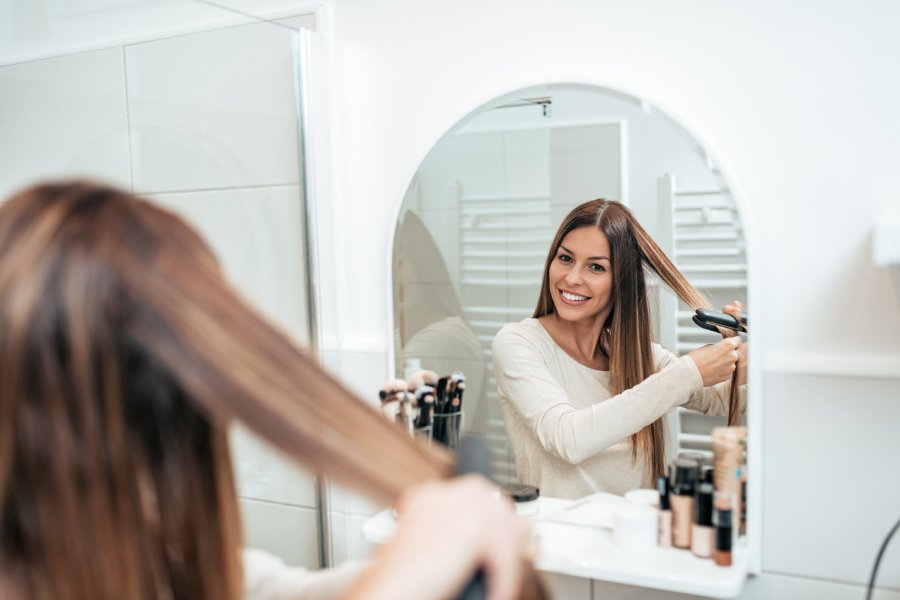 A woman with brown and blonde highlighted hair is smiling is straightening her hair in a salon chair.