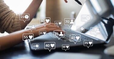 A woman in front of her laptop. Digital speech bubbles with the symbols for followers, likes, and comments fill the foreground.