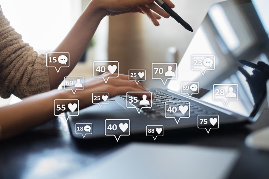 A woman in front of her laptop. Digital speech bubbles with the symbols for followers, likes, and comments fill the foreground.
