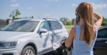 A woman stands on the street while talking on the phone and looking at a white car that has been wrecked into.