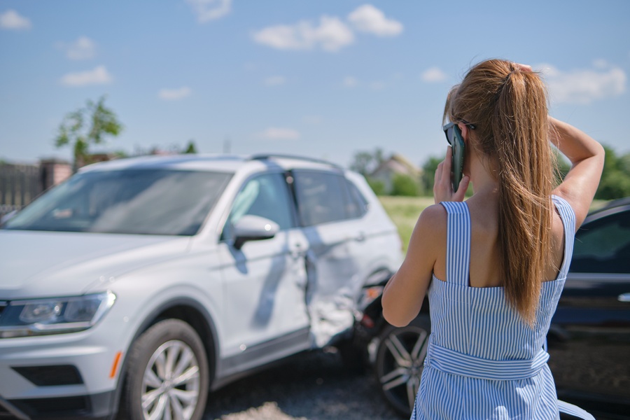 A woman stands on the street while talking on the phone and looking at a white car that has been wrecked into.