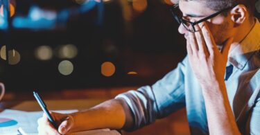 A young man works in an office with very poor lighting. He writes on a notepad and tiredly rubs his eyes.
