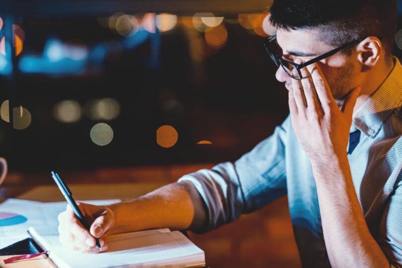 A young man works in an office with very poor lighting. He writes on a notepad and tiredly rubs his eyes.