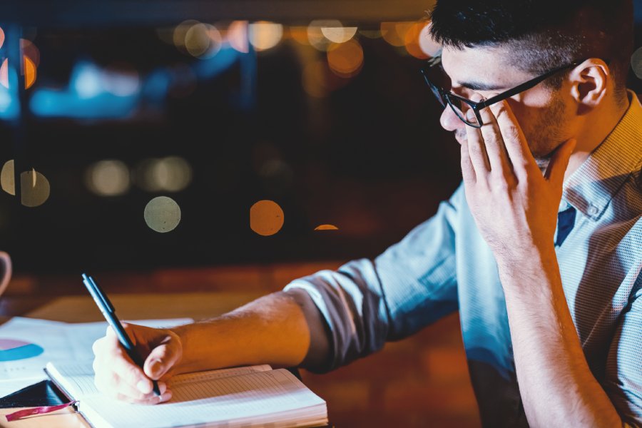 A young man works in an office with very poor lighting. He writes on a notepad and tiredly rubs his eyes.
