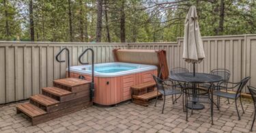 A brown hot tub sits on a brick patio next to a wrought-iron table and chairs. Wooden stairs lead to the open hot tub.
