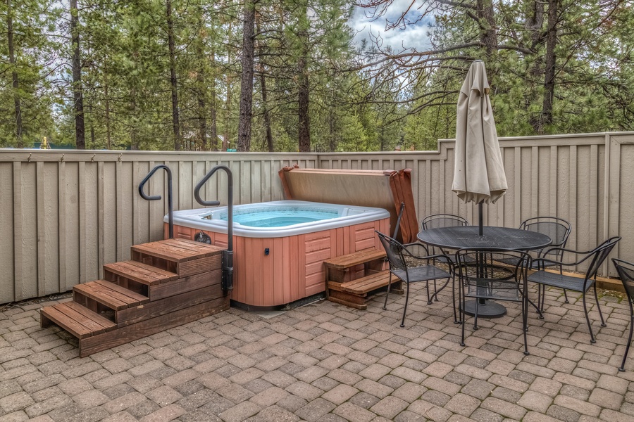 A brown hot tub sits on a brick patio next to a wrought-iron table and chairs. Wooden stairs lead to the open hot tub.