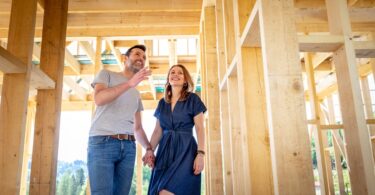 A man and woman hold hands inside a home under construction with wooden beams for the walls and roof.
