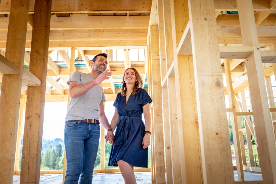 A man and woman hold hands inside a home under construction with wooden beams for the walls and roof.