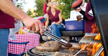 A man using silver tongs to pick up a sausage on a grill. Next to him are men and women grouped around a cooler.
