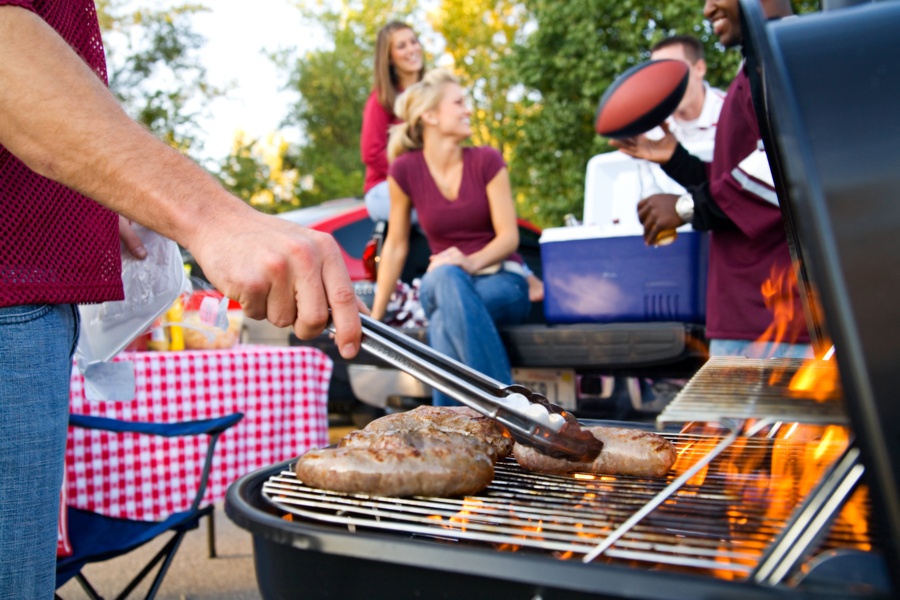 A man using silver tongs to pick up a sausage on a grill. Next to him are men and women grouped around a cooler.
