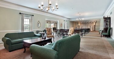 The large viewing room of a funeral home, including rows of chairs, two couches, chandeliers, and a coffee table.
