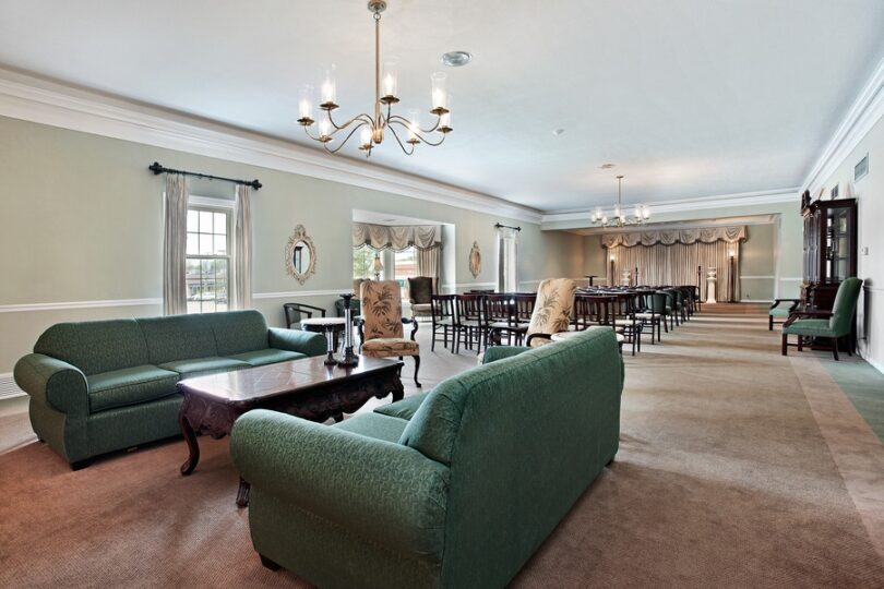 The large viewing room of a funeral home, including rows of chairs, two couches, chandeliers, and a coffee table.