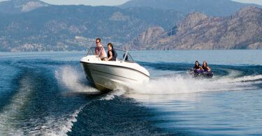 A motorboat on the water pulling two people on an inner tube over the wake with mountains in the background.