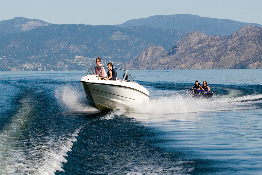 A motorboat on the water pulling two people on an inner tube over the wake with mountains in the background.