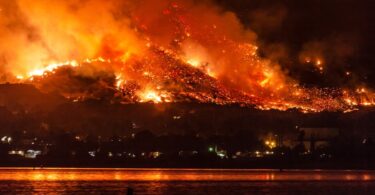 A massive wildfire spreading across a mountain of trees. There are homes at the base of the mountain near a lake.