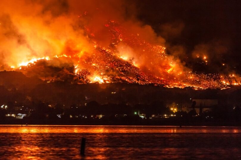 A massive wildfire spreading across a mountain of trees. There are homes at the base of the mountain near a lake.