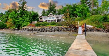 A luxury waterfront cottage on the edge of a lake with a forest in the background. There is a wooden dock on the shore.