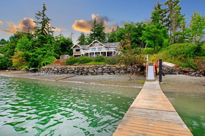 A luxury waterfront cottage on the edge of a lake with a forest in the background. There is a wooden dock on the shore.