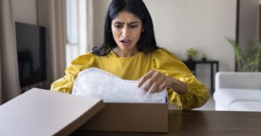 A female customer sitting at her dining room table opening a package with a shocked and disappointed facial expression.