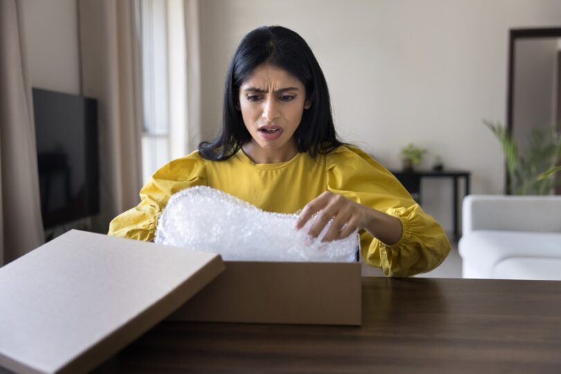 A female customer sitting at her dining room table opening a package with a shocked and disappointed facial expression.